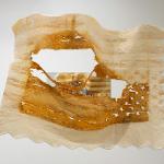 Wall-mounted mixed-media artwork on translucent, irregularly edged fabric, stained in warm amber and tan tones, with a large central cutout revealing layered imagery behind it; fragments of an American flag pattern, including stripes and white stars, are embedded within the material.