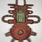 Wall-mounted sculptural form with a central seated figure, radiating extensions and circular motifs, finished in rust red and turquoise tones against a white background.