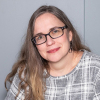 Heather facing forward smiling, wearing glasses, long brown hair, gray and white plaid shirt