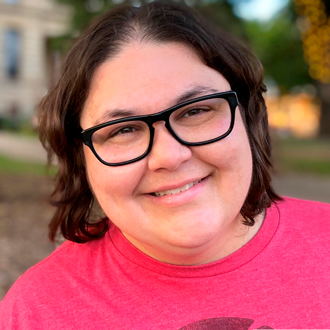 Jenn is facing forward and smiling. She has brown, shoulder-length hair, wears black glasses and a hot pink T-shirt.