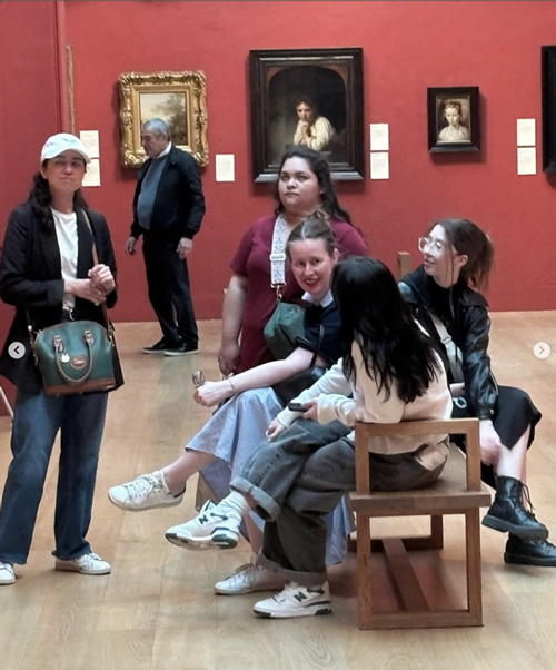 Students standing with others sitting on a bench inside an art museum.