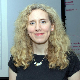 Carey is facing forward and smiling. She has long blonde, wavy hair and wears a black top.