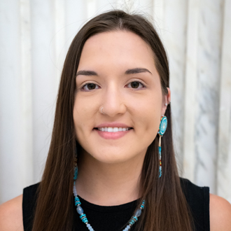 Katherine is facing forward and smiling. She has long dark hair and wears turquoise earrings, a necklace and a black top.