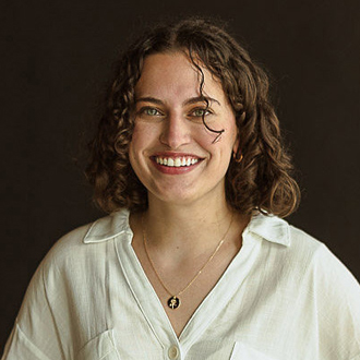 Gina is facing forward and smiling. She has brown, curly hair and wears a white top.