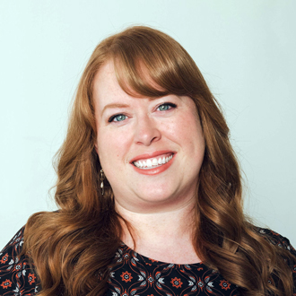 Madeleine is facing forward and smiling. She has long red hair and wears a brown floral printed top.