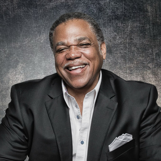 Terence is facing forward and smiling. His hands are on a desk. He wears a dark suit and white shirt.