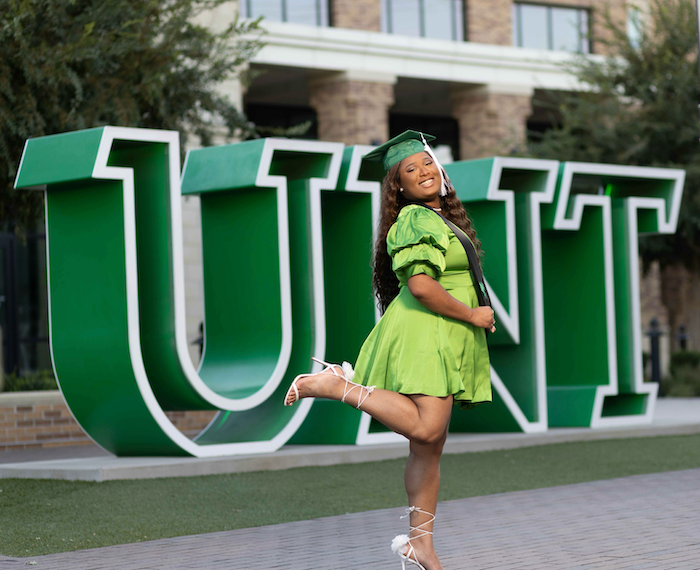 Shanice is in front of the UNT Welcome Center sign in a green dress and kicking up her foot in celebration.