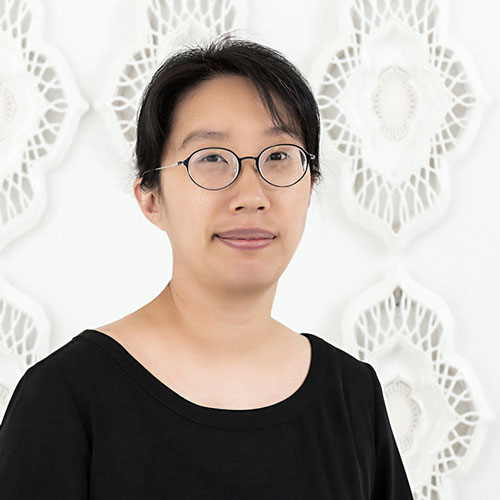 Eliza Au facing forward. She has dark hair, wears glasses and a black scoop-neck top against a white background.