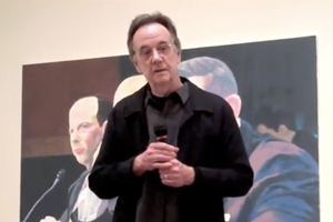 Ed Blackburn is standing, holding a microphone with paintings behind him.