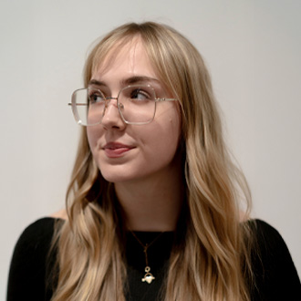 Rachel is looking up to her right. She has long blonde hair and wears glasses.