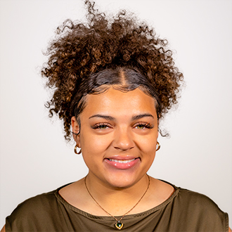 Lauryn is facing forward and smiling. Her brown hair is in an updo and her top is brown,