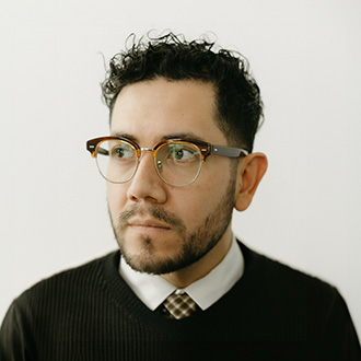 Daniel Mejia is looking to the right. He has short dark hair, wears glasses, a plaid tie, a white shirt and dark sweater.