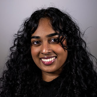 Sriya is facing forward and smiling. She has dark curly hair and wears a dark top.