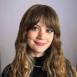 Brennah is facing forward and smiling. She has long brown, wavy hair and wears a black turtleneck top.