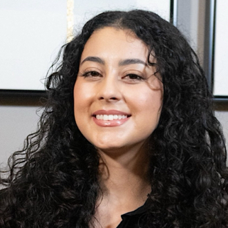 Laura is facing forward and smiling. She has long, dark hair in ringlets and wears a dark top.