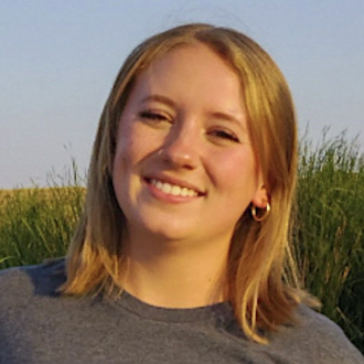 Sydnie is facing forward and smiling. She has blonde hair and wears a gray top.