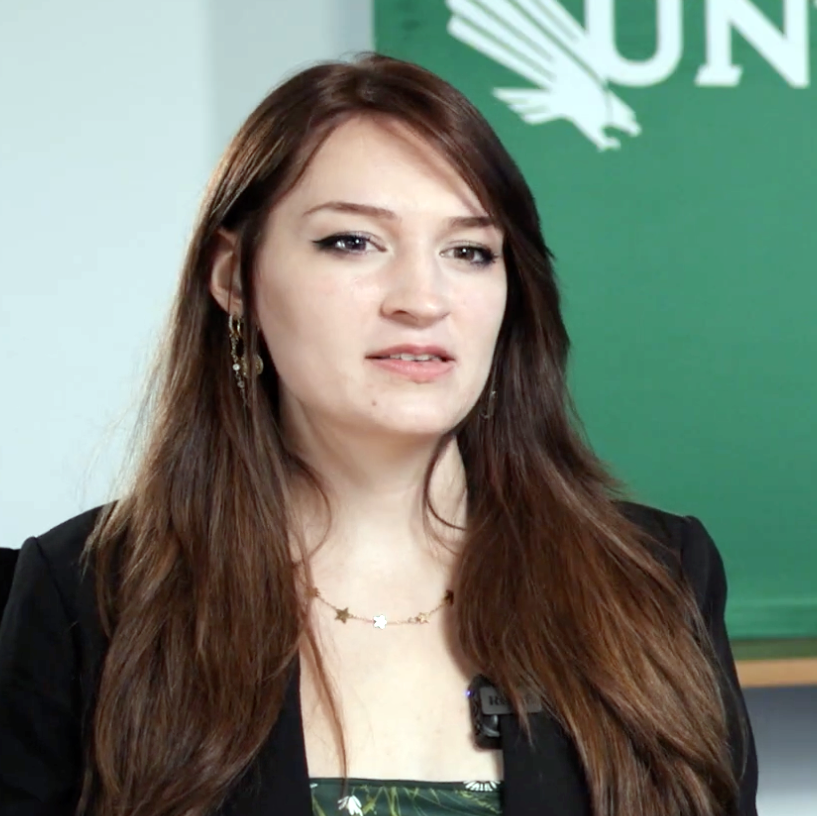 Natasha Sturdevant is looking toward her left. She has long brown hair, wears an necklace and a black jacket.