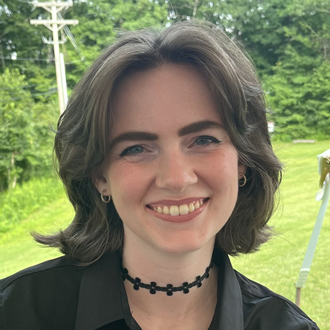 Emily facing forward and smiling. She has short brown hair and trees in the background.