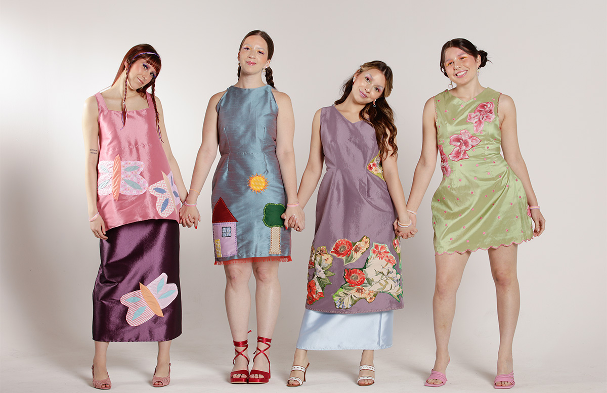 Four ladies stand together holding hands wearing the colorful designs. The first model is wearing a pink top and a long purple skirt both with patchwork graphics of some butterflies. The second model is wearing a blue dress with graphics of a house, a tree and the sun. The third model is wearing a lilac dress with a light blue skirt underneath. The dress has some yellow, red and blue flowers stitched on. The fourth model is wearing a green dress with pink polkadots and a pink trim at the hem. The dress also has some pink flowers stitched on.  
