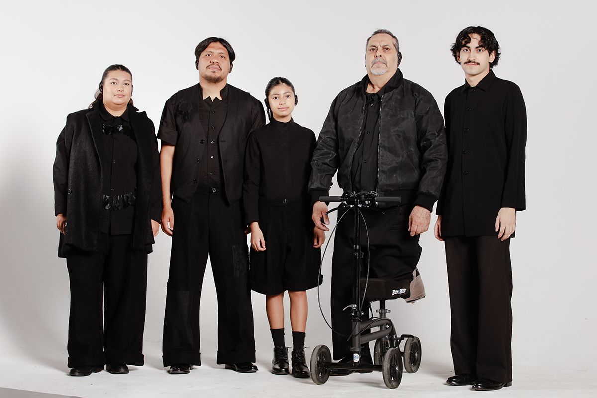 Four models and the designer, all wearing full black outfits, varying in age and silhouette.
