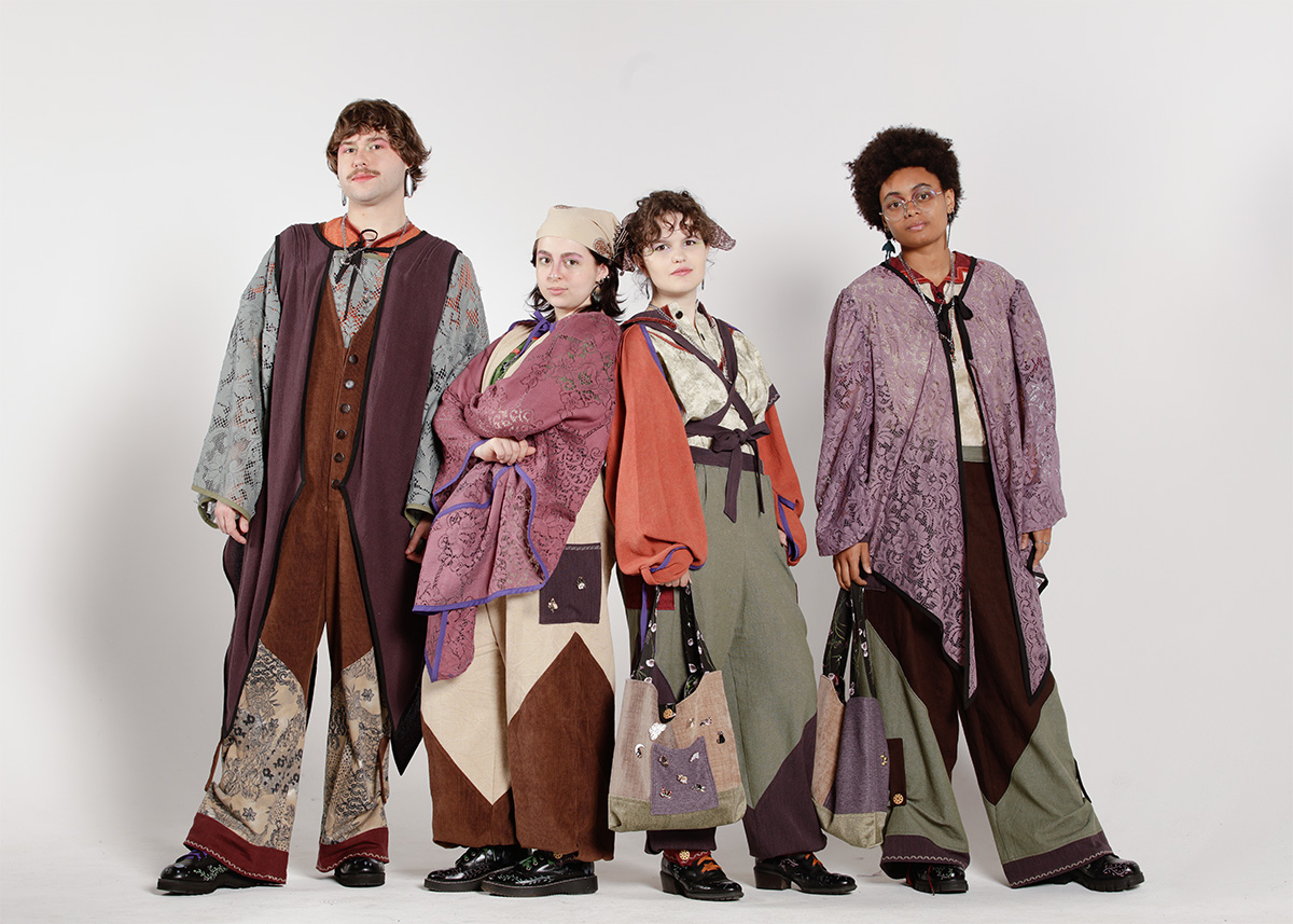 Four models wearing multi-layered garments in tones of green, brown, purple and rusty orange standing in front of a white studio backdrop.