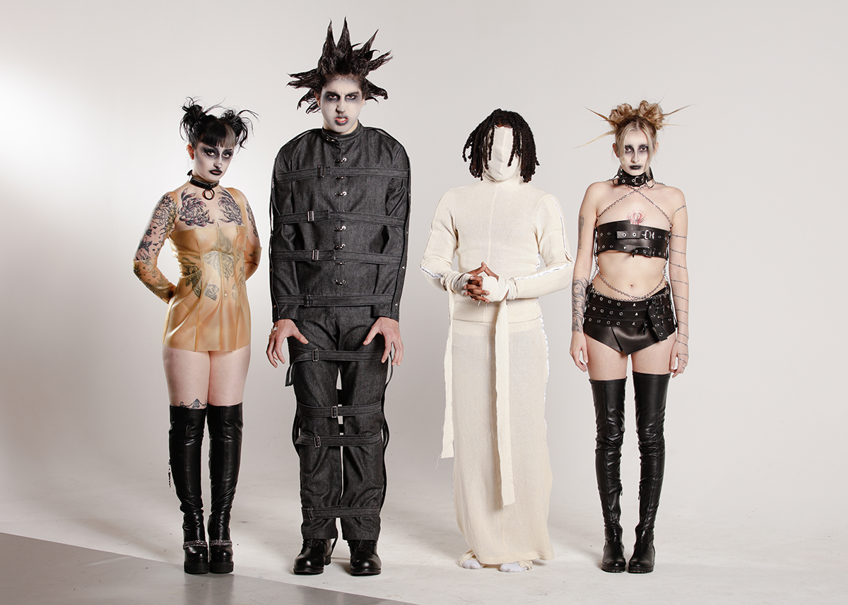 Four models (left to right), first female model posing in transparent leather corset with sleeves, second male model posing in black selvedge denim jacket and pants, third male model posing in gauze mask, long sleeve, and skirt, fourth female model posing in black leather neck, bust, and hip belts connected via chains. 