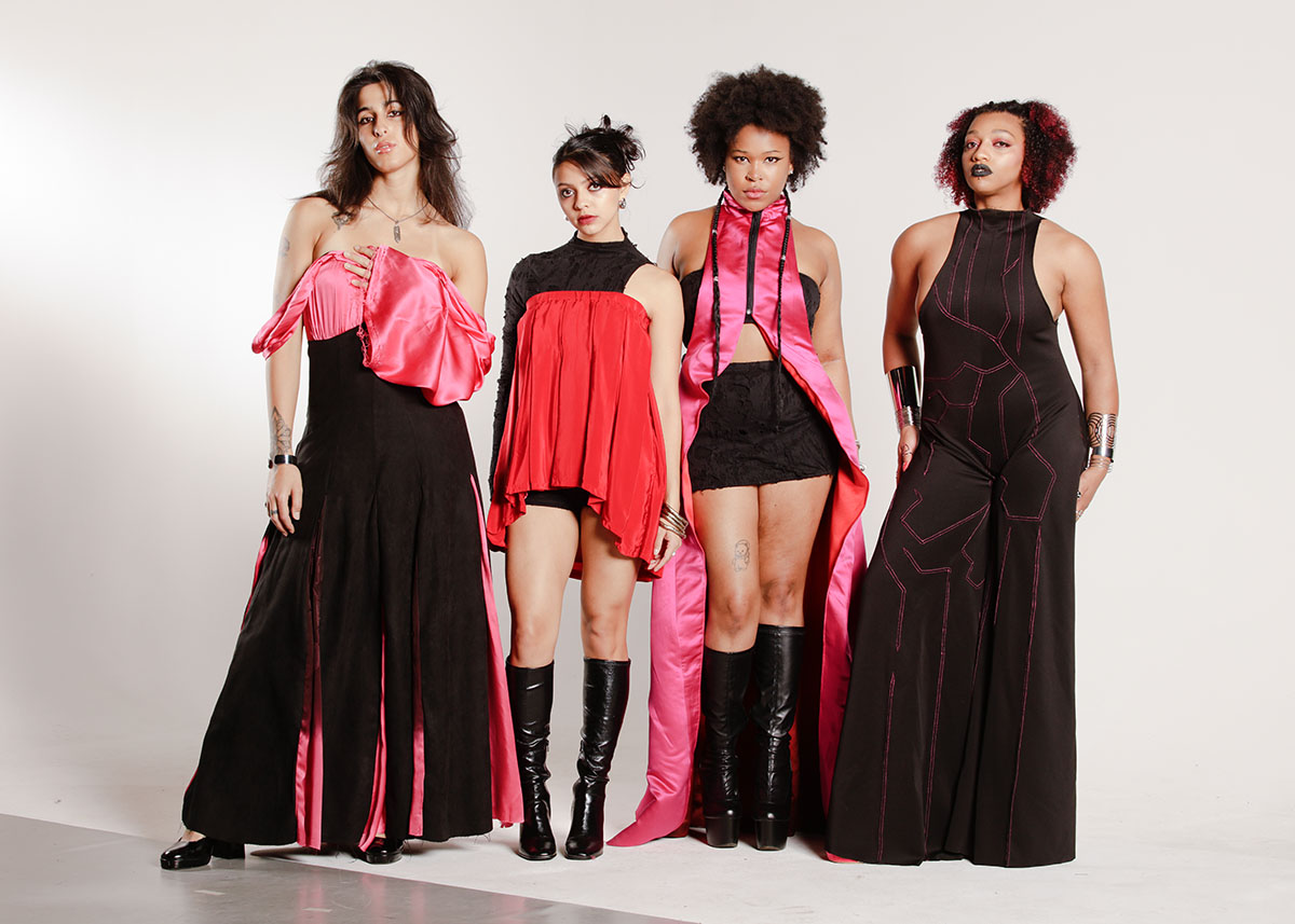 Four models standing together in black, pink, and red garments made of satin, silk, suede, and distressed knit, and spandex.