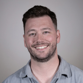 Drew is facing forward and smiling. He has short brown hair, a beard and wears a blue shirt.