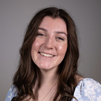 Kate is facing forward and smiling. She has long brown hair and wears a blue top.