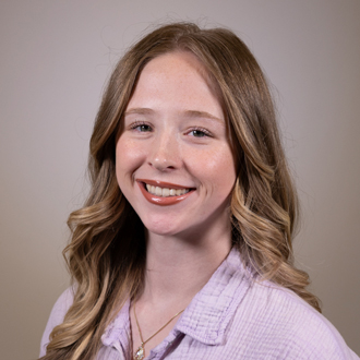 Meredith is facing forward and smiling. She has long blonde hair and wears a pink shirt.