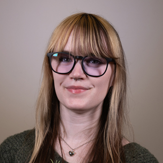 Hanna is facing forward and smiling. She has brown and blonde hair with bangs and wears glasses.