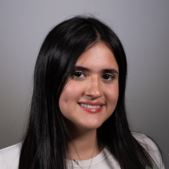 Cynthia is facing forward and smiling. She has long dark hair and wears a white top.
