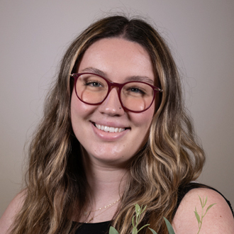 Indi is facing forward and smiling. She has long blonde hair and wears glasses.