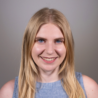 Phoebe is facing forward and smiling. She has long, blonde hair and wears a blue top.