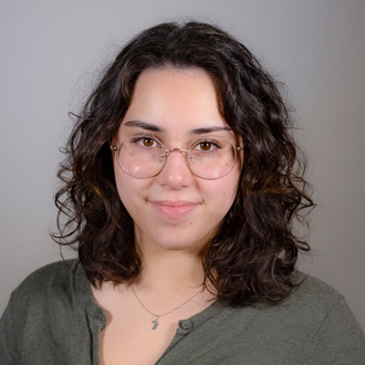 Sophia is facing forward. She has shoulder-lenght brown, wavy hair and wears glasses.