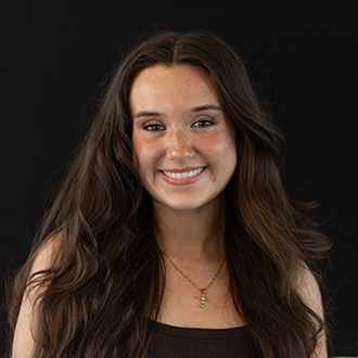 Trinity is facing forward and smiling. She has long wavy, dark hair and wears a black top.