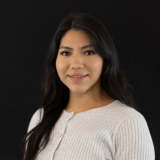 Gloria is facing forward and smiling. She has long wavy, dark hair and wears a white top.