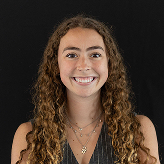 Bailee is facing forward and smiling. She has long curly, brown hair and wears a black top.