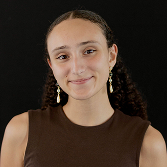 Hannah is facing forward and smiling. She has long, curly brown hair and wears a brown top.