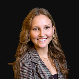 Celia is facing forward and smiling. She has wavy light-brown hair and is wearing a black-and-tan checked blazer.