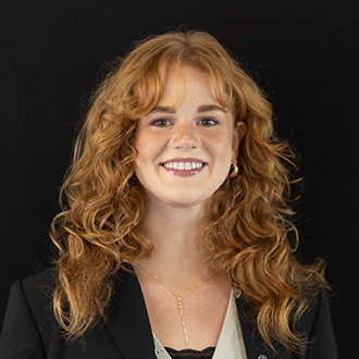 Bethany is facing forward and smiling. She has long wavy, red hair and wears a black top.