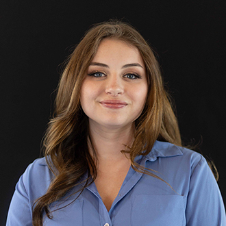 Natalie is facing forward and smiling. She has long brown hair and wears a blue top.