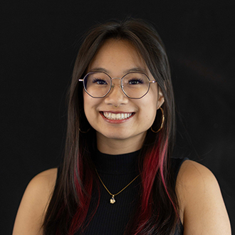 Lyna is facing forward and smiling. She has long dark hair and wears a black top.