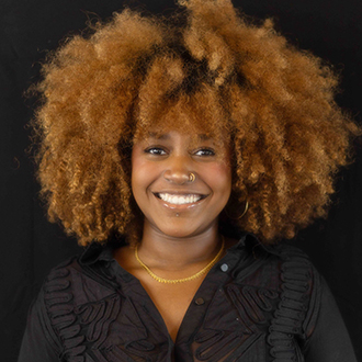 Laila is facing forward and smiling. She has medium-length, curly brown hair and wears a black top.