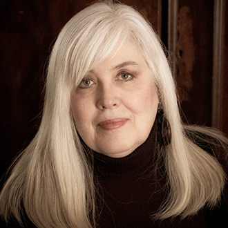 Karen is facing forward and smiling. She has long blonde hair and wears a black turtle-neck top.