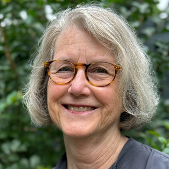 Dornith is facing forward and smiling. She wears a short bob, brown glasses and a gray shirt.