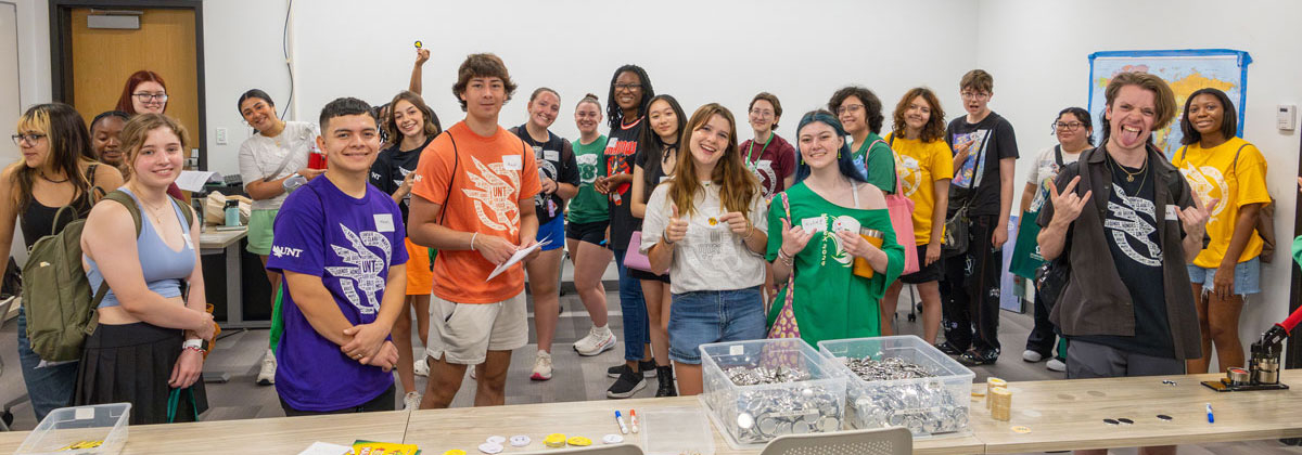 First Flight 2024: about 20 students smiling at the camera as they begin a button-making activity