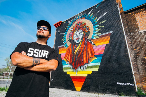 Victor Quinonez is standing next to a large art exhibit of a yellow popsicle wearing a dark cap, T-shirt and jeans. 