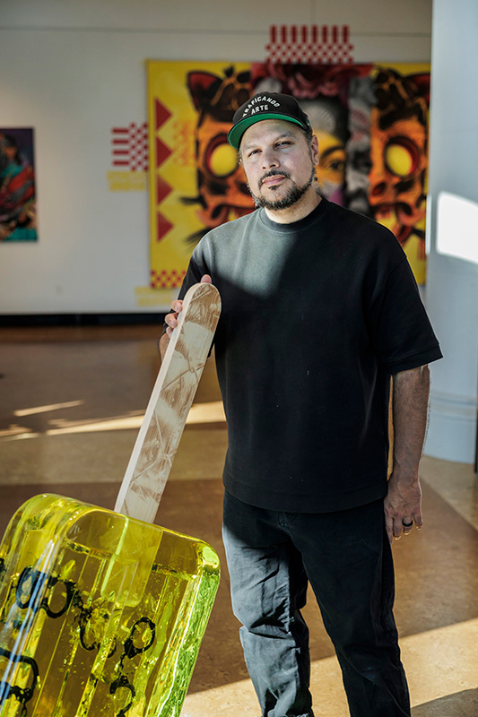 Victor Quinonez is standing next to a large art exhibit of a yellow popsicle wearing a dark cap, T-shirt and jeans. 