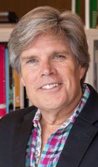 Michael is facing forward and smiling. He has light brown hair and wears a plaid shirt and navy jacket.